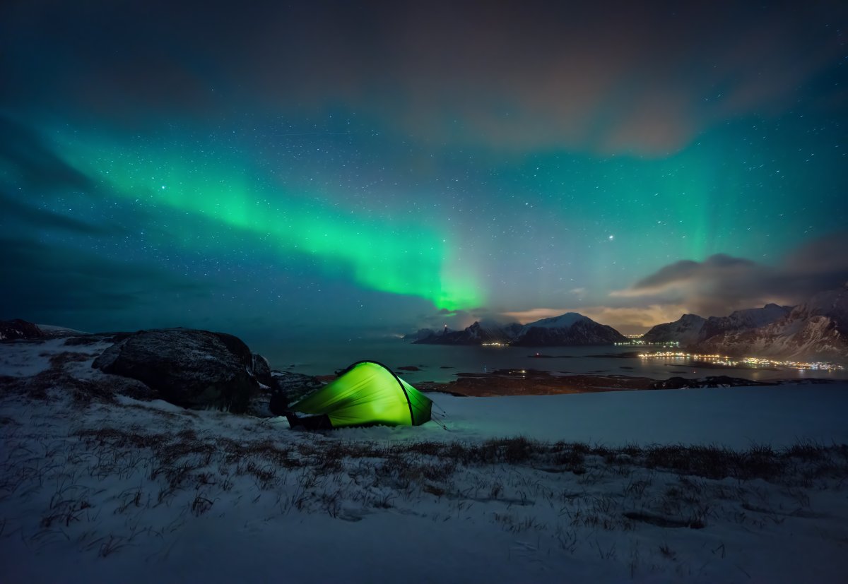 Nuit sous les aurores boréales en Norvège – Émerveillement polaire en bivouac Tente de camping illuminée sous des aurores boréales vertes dans un paysage enneigé de Norvège, avec vue sur les montagnes et un fjord nocturne scintillant.