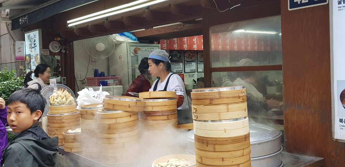 Scène de rue en Corée : préparation de dumplings dans un marché traditionnel Femme coréenne cuisinant des dumplings à la vapeur dans un marché de rue animé, avec des paniers en bambou fumants et des passants en arrière-plan