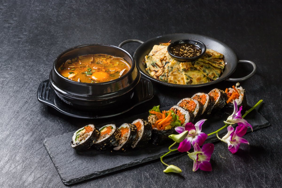 Gimbap, soupe épicée et pancakes coréens – Cuisine de Corée du Sud Gimbap roulé coréen accompagné de soupe au tofu épicée et de pancakes aux légumes, présentés sur une table noire avec fleurs décoratives.