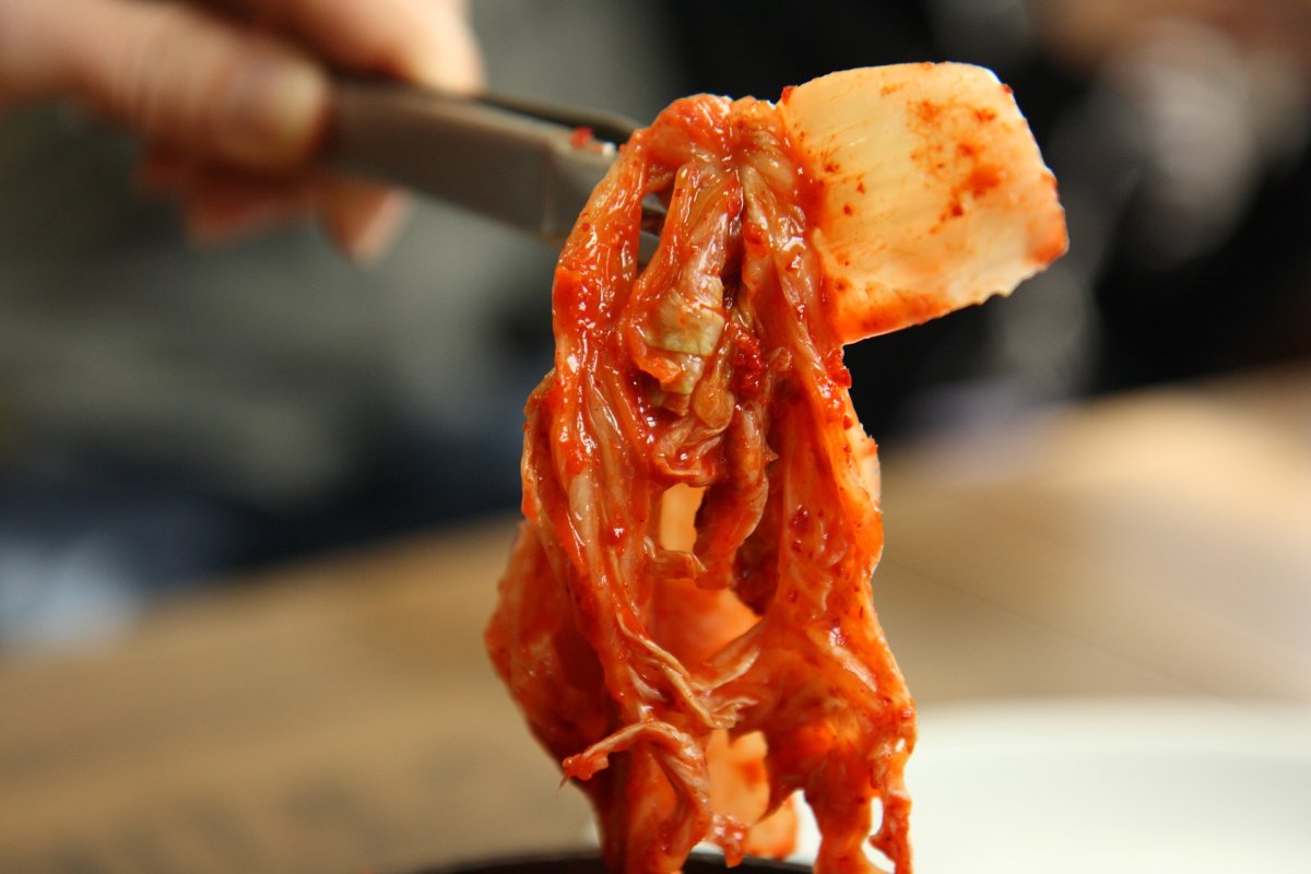 Une bouchée de caractère Un gros plan sur des baguettes saisissant un morceau de kimchi bien fermenté. La texture brillante, les fibres du chou et la couleur vive du piment révèlent toute la puissance aromatique de ce plat emblématique. Cette image illustre parfaitement l’intensité gustative du kimchi et son rôle essentiel à table.