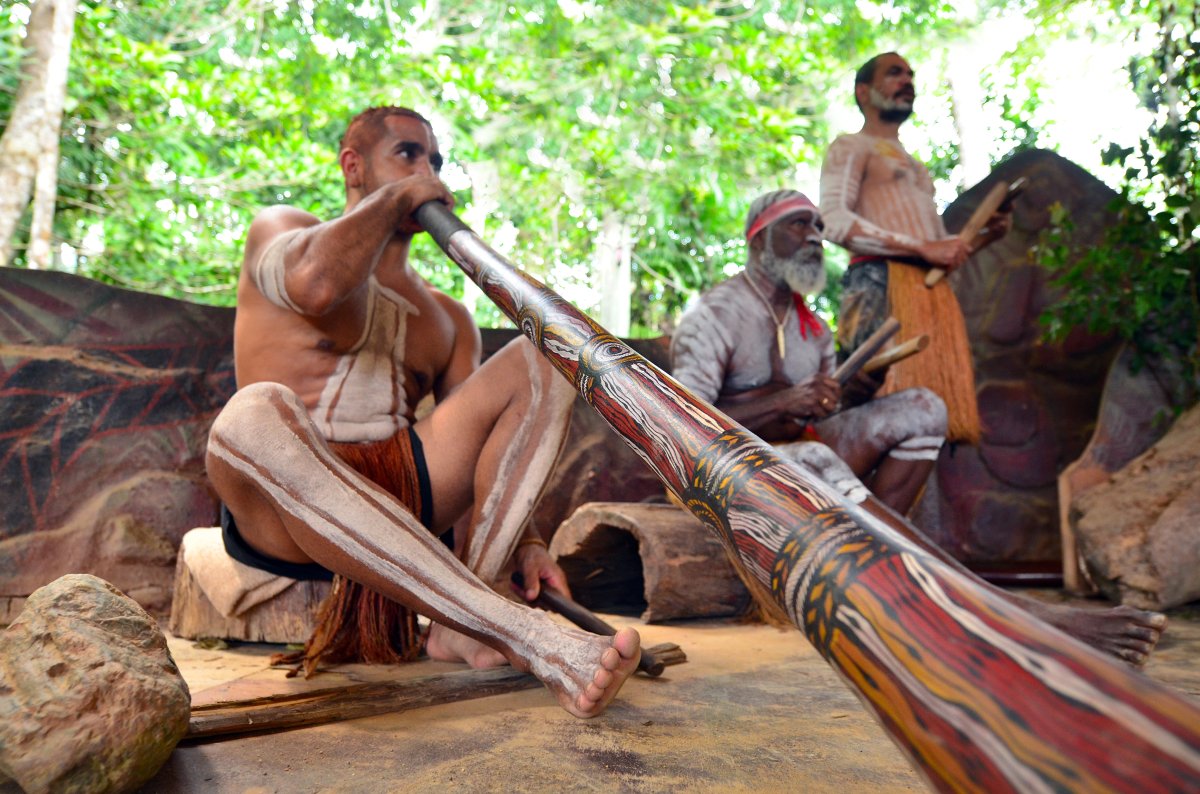 Musique aborigène – Rituel ancestral au didgeridoo Homme aborigène jouant du didgeridoo lors d’un rituel traditionnel, entouré de musiciens en tenue cérémonielle, illustrant la richesse culturelle et spirituelle des peuples premiers d’Australie.