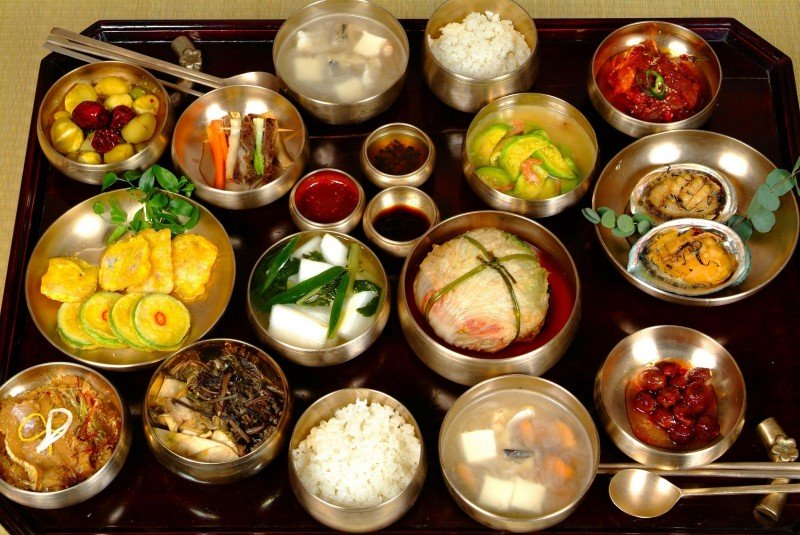 L’abondance harmonieuse d’un repas coréen Cette vue d'ensemble montre une table typique de restaurant coréen : une grande corbeille de banchan au centre, accompagnée de plats chauds, de soupes et d’une marmite de riz. On y aperçoit du kimchi, mais en tant qu’un élément parmi d'autres. Idéale pour illustrer l’aspect rituel et collectif du repas.