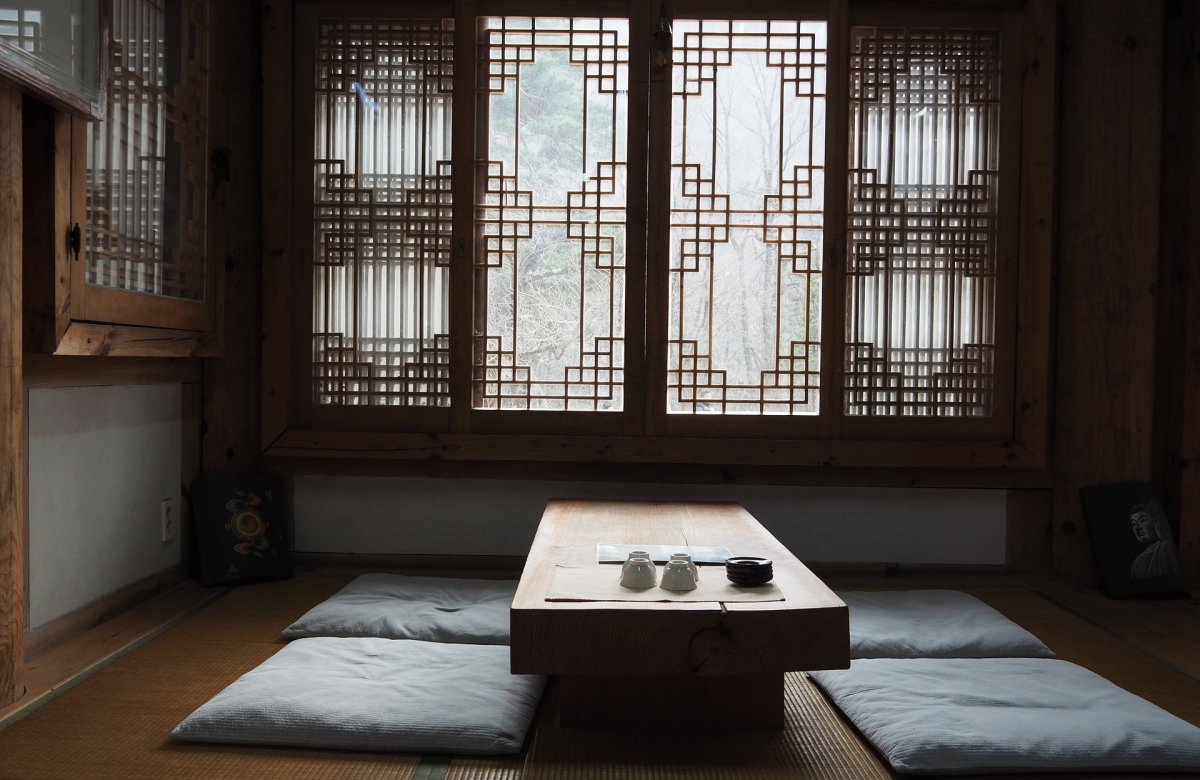 L’élégance sobre du hanok Une table basse en bois brut, posée sur un sol en tatami, entourée de coussins, baignée d’une lumière douce filtrée par des fenêtres à motifs géométriques traditionnels. Cette pièce évoque la sérénité, le respect des gestes simples, et la place centrale du repas dans l’art de vivre coréen.